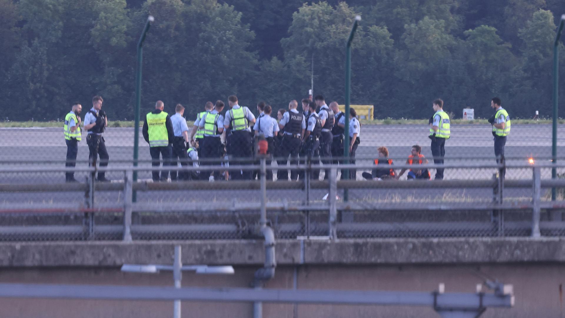 Das Foto wurde aus großer Entfernung aufgenommen. Zu sehen sind viele Polizisten, die in roten Warnwesten hinter einem Zaun um die abgeklebten Aktivisten stehen.