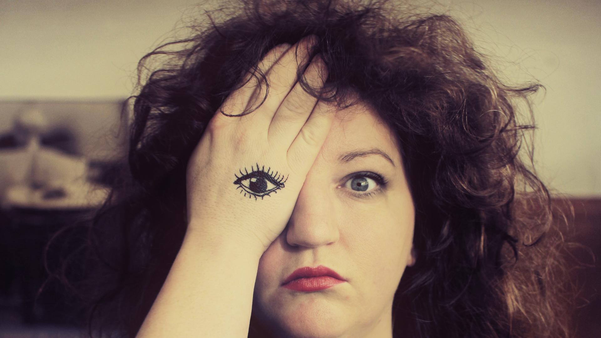 Musikerin Anna Mateur im Porträt. Ihre rechte Hand verdeckt ihr rechtes Auge. Auf ihre Handfläche ist ein Auge gemalt.
