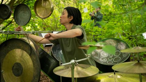 Marie Watanabe spielt unter einem günen Blätterdach Schlagzeug
