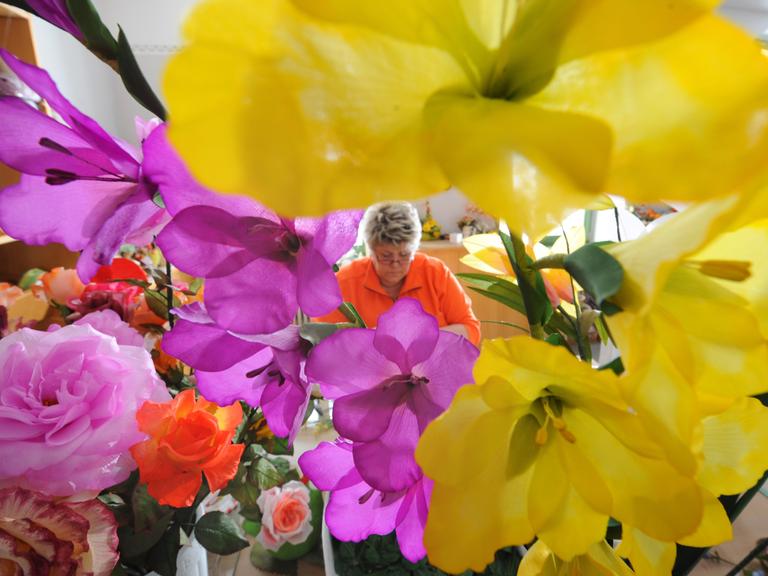 Im Vordergrund sind in der Schauwerkstatt "Deutsche Kunstblume Sebnitz" üppige, verschiedenfarbige Kunstblumen zu sehen, im Hintergrund eine Frau, die weitere Blumen fertigt.