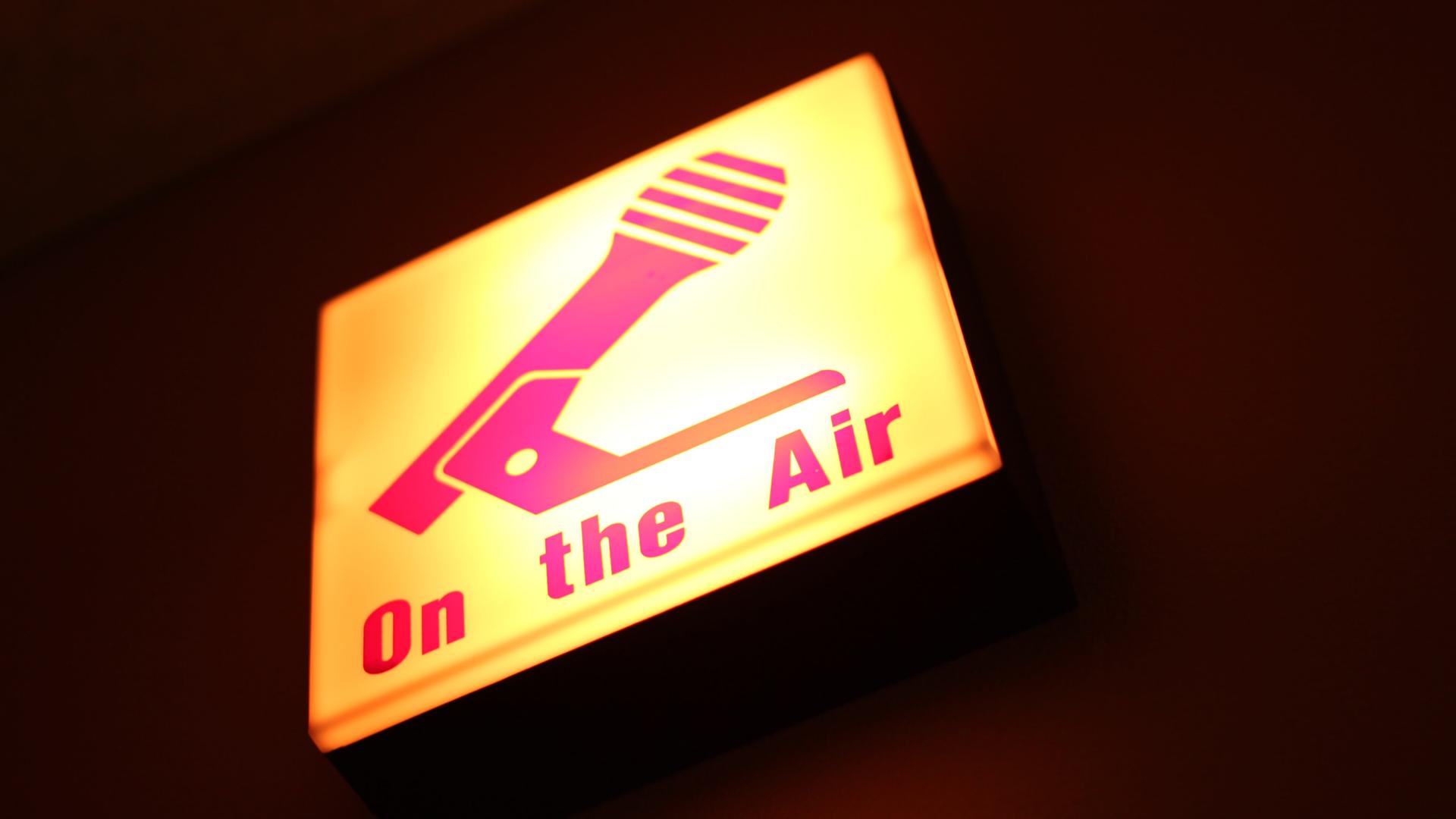 Gelblich-pink leuchtendes Schild mit der Aufschrift "on the Air" und einem Mikrofon Gelblich-pink leuchtendes Schild mit der Aufschrift "on the Air" und einem Mikrofon