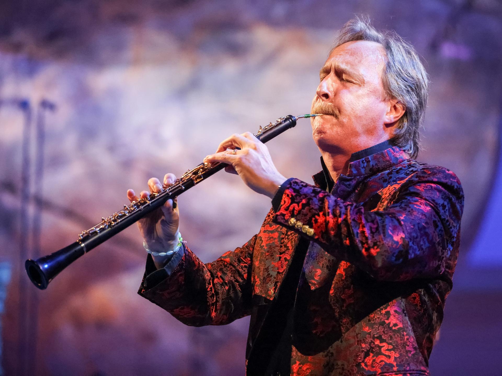 Ein Mann mit graumeliertem Haar spielt eine Oboe. Er trägt einen Oberlippenbart, sein Jackett erscheint in bunten Farben. Der Mann, Albrecht Mayer, ist im Profil zu sehen, er hat beim Spiel die Augen geschlossen. 