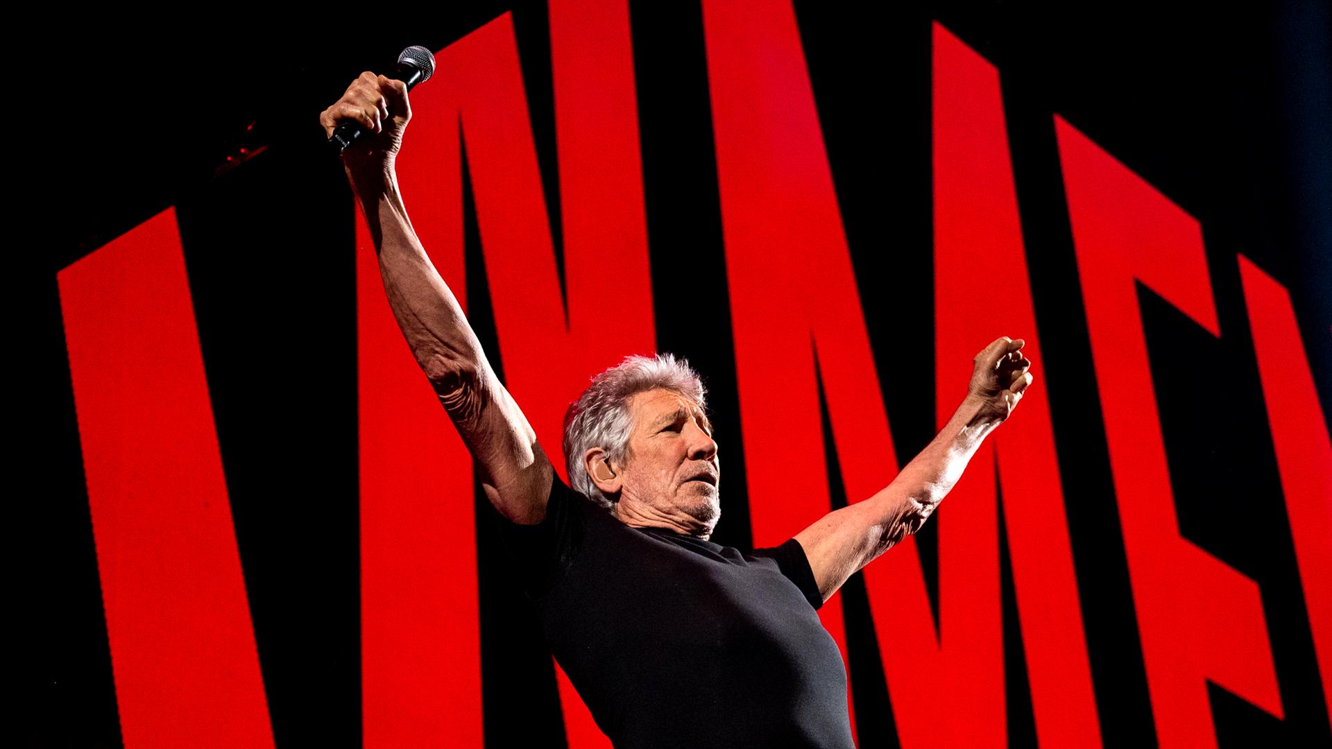 Roger Waters auf der Bühne in Hamburg mit ausgebreiteten Armen. Hinter ihm rote Symbole vor schwarzem Hintergrund. Roger Waters auf der Bühne in Hamburg mit ausgebreiteten Armen. Hinter ihm rote Symbole vor schwarzem Hintergrund.