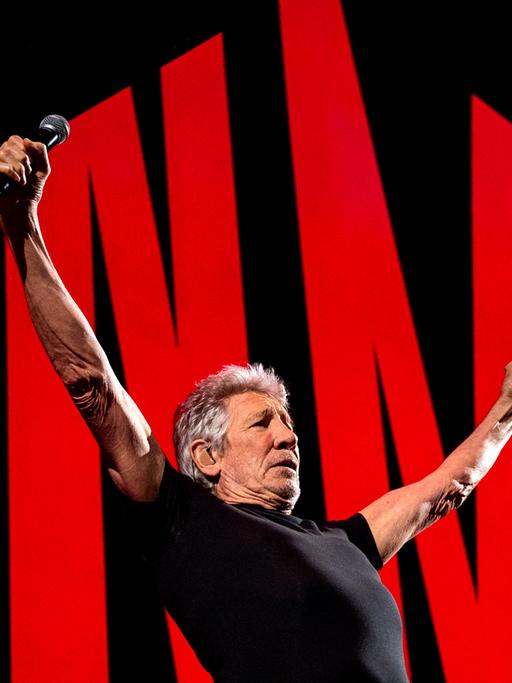 Roger Waters auf der Bühne in Hamburg mit ausgebreiteten Armen. Hinter ihm rote Symbole vor schwarzem Hintergrund.