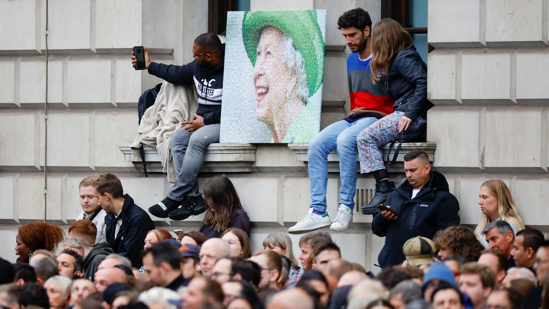 Zum Staatsbegräbnis von Queen Elizabeth II. drängen sich Menschen vor einem Gebäude, einige sitzen auf Fensterbänken, zwischen sich ein etwa 1x1 Meter großes Poprträt der Königin.