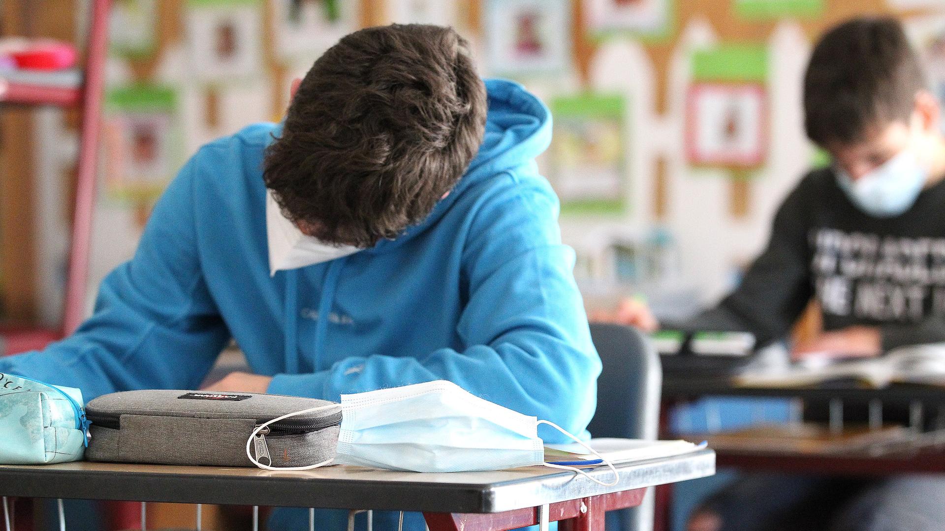 Zwei Schüler mit Masken sitzen in einem Klassenraum und schreiben an ihrem Pult.