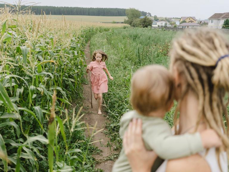Ein Mädchen läuft auf einem Maisfeld seiner Mutter entgegen, im Hintergrund ist ein Dorf zu sehen.