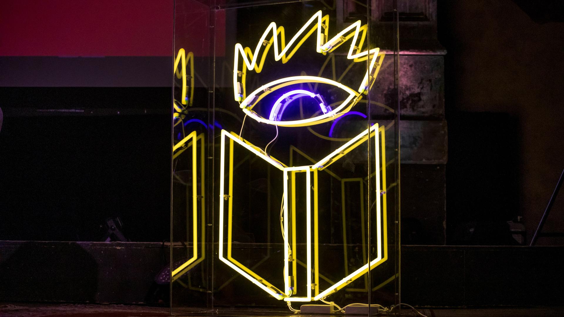 Gelbe, weiße und blaue Neonröhren bilden das Logo der Buchmesse Leipzig, ein weit geöffnetes Auge über einem aufgeschlagenen Buch. Gelbe, weiße und blaue Neonröhren bilden das Logo der Buchmesse Leipzig, ein weit geöffnetes Auge über einem aufgeschlagenen Buch.