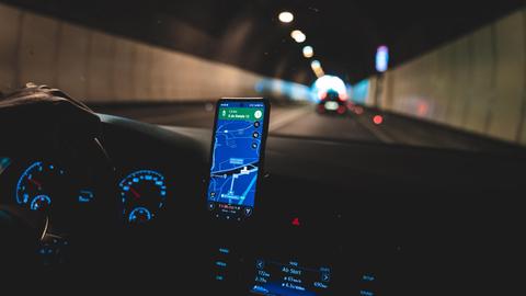 Ein Wagen fährt im Dunkeln durch einen Tunnel, im Cockpit des Fahrzeugs leuchten Navigation diverse Lichter sowie ein Smartphone mit der Google Maps App. 