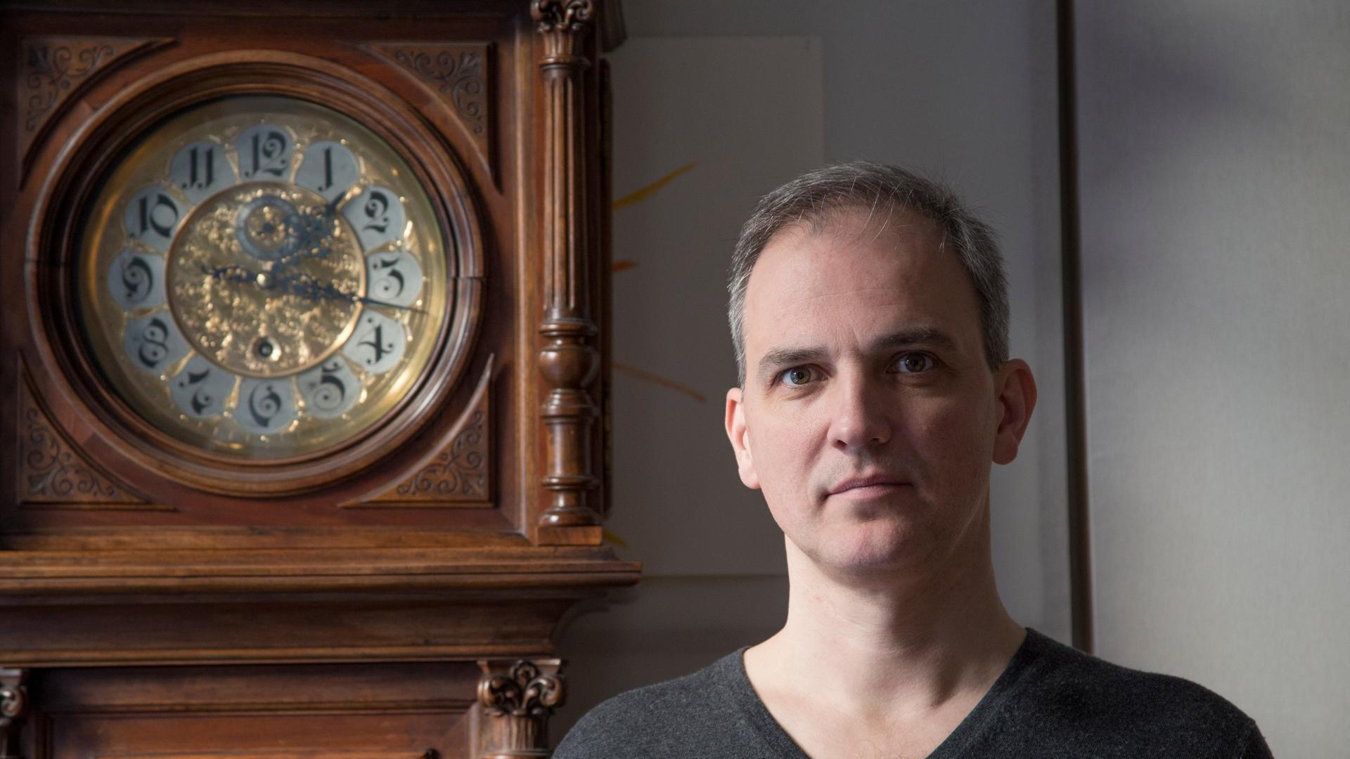Volkmar Klien schaut neben dem goldverzierten Ziffernblatt einer neugotischen Gründerzeit-Standuhr im die Kamera Volkmar Klien schaut neben dem goldverzierten Ziffernblatt einer neugotischen Gründerzeit-Standuhr im die Kamera