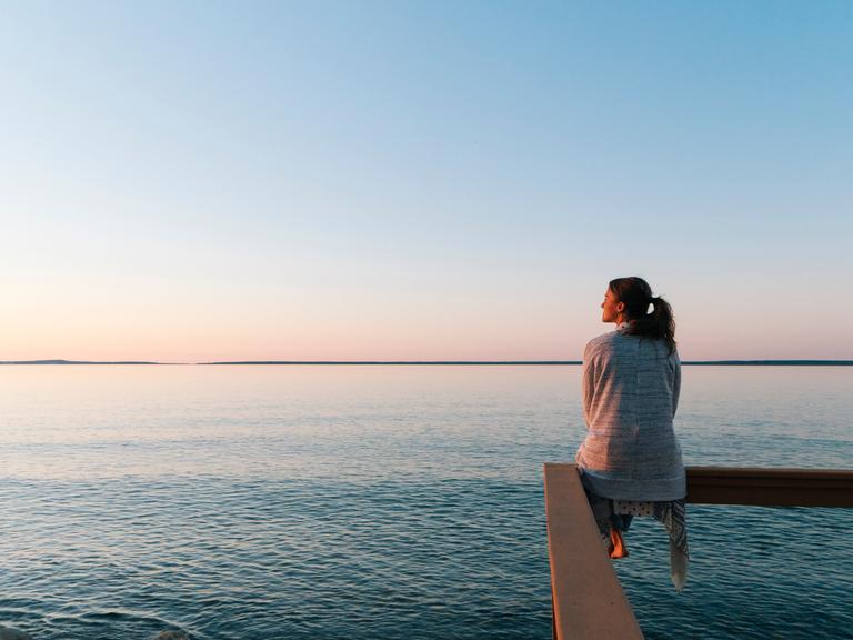 Eine junge Frau sitzt auf einem Geländer an einem See und schaut in die Weite.