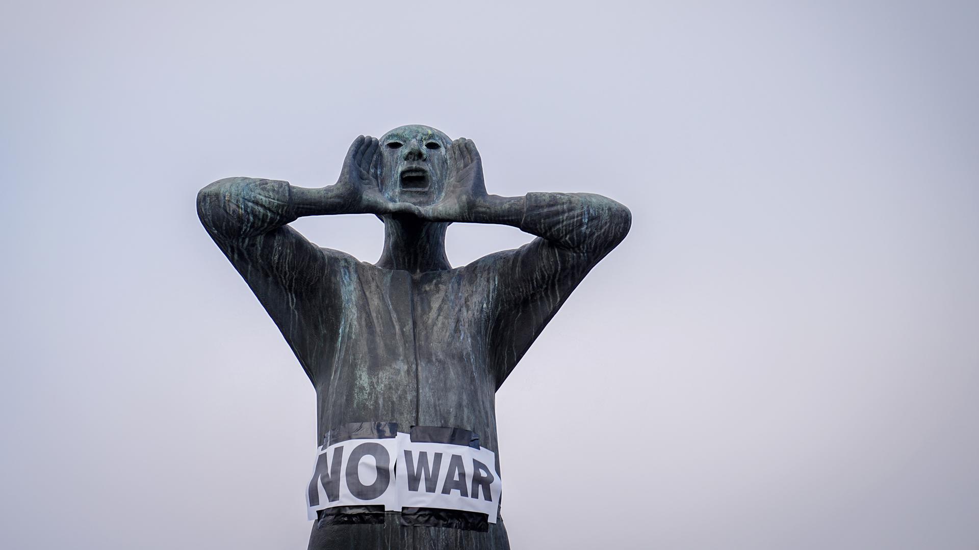 Plakate mit der Aufschrift "No war" kleben an einer männlichen Bronzefigur, deren Händen am weit geöffenet Mund plaziert sind.