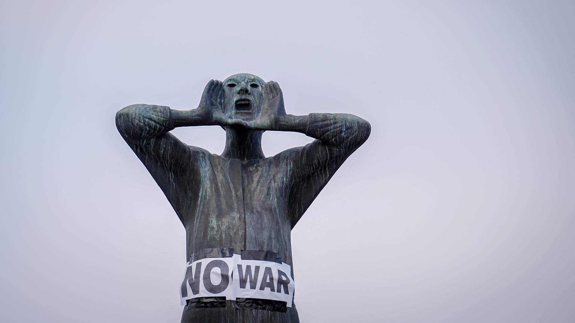 Plakate mit der Aufschrift "No war" kleben an einer männlichen Bronzefigur, deren Händen am weit geöffenet Mund plaziert sind. Plakate mit der Aufschrift "No war" kleben an einer männlichen Bronzefigur, deren Händen am weit geöffenet Mund plaziert sind.