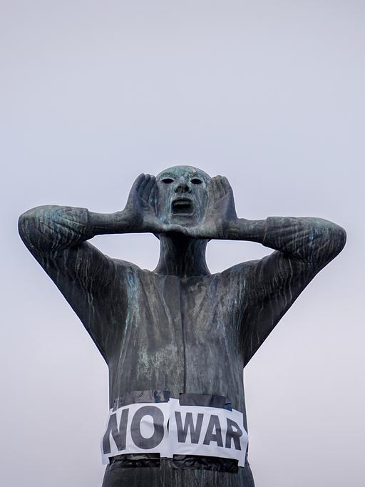 Plakate mit der Aufschrift "No war" kleben an einer männlichen Bronzefigur, deren Händen am weit geöffenet Mund plaziert sind.