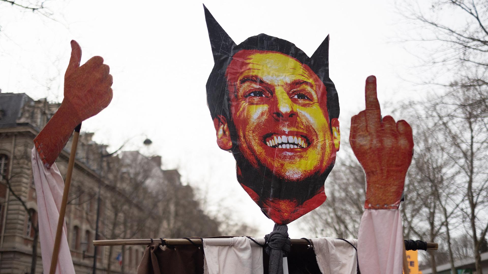 Protestler halten ein Schild hoch, das Frankreichs Präsident Macron mit Teufelshörnern und ausgestrecktem Mittelfinger zeigt Protestler halten ein Schild hoch, das Frankreichs Präsident Macron mit Teufelshörnern und ausgestrecktem Mittelfinger zeigt