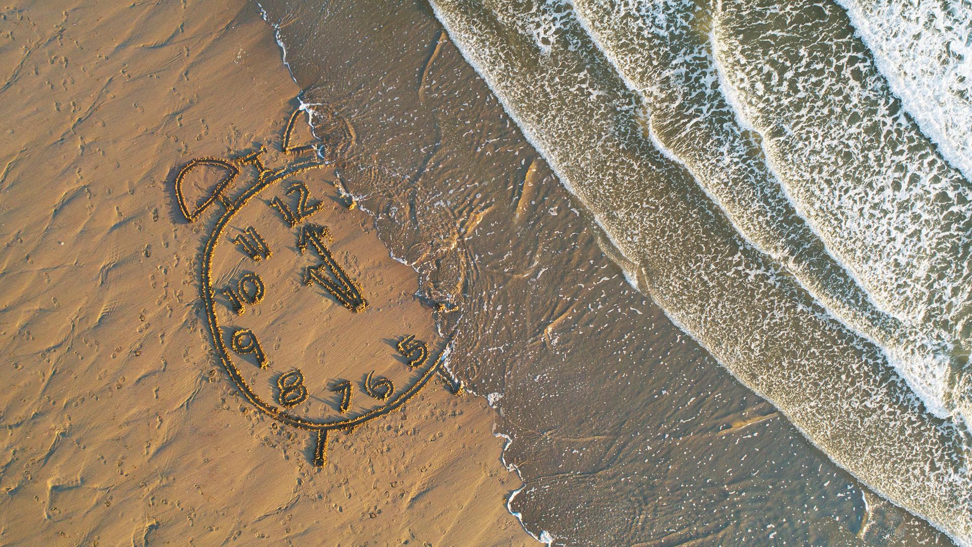 Am Strand: Eine in den Sand gemalte Uhr wird vom Meer überflutet.