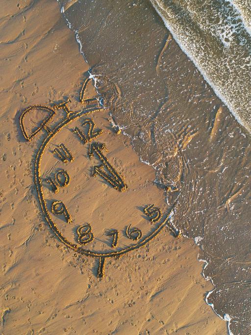 Am Strand: Eine in den Sand gemalte Uhr wird vom Meer überflutet.