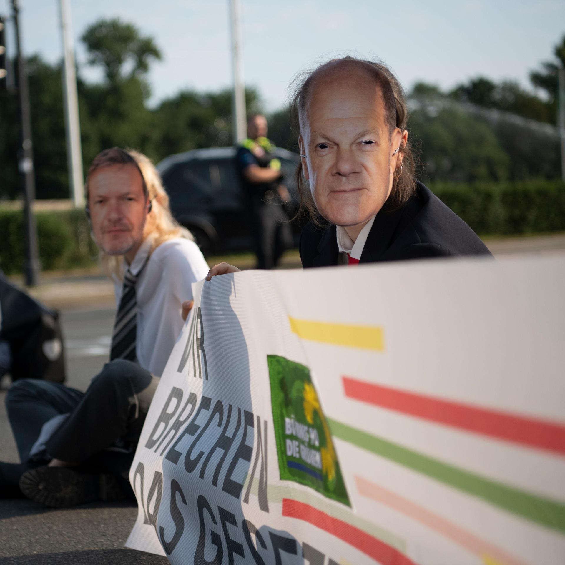 Zwei Demonstranten sitzen auf der Straße. Sie tragen Masken mit Fotos von Kanzler Scholz und Wirtschaftsminister Habeck. 