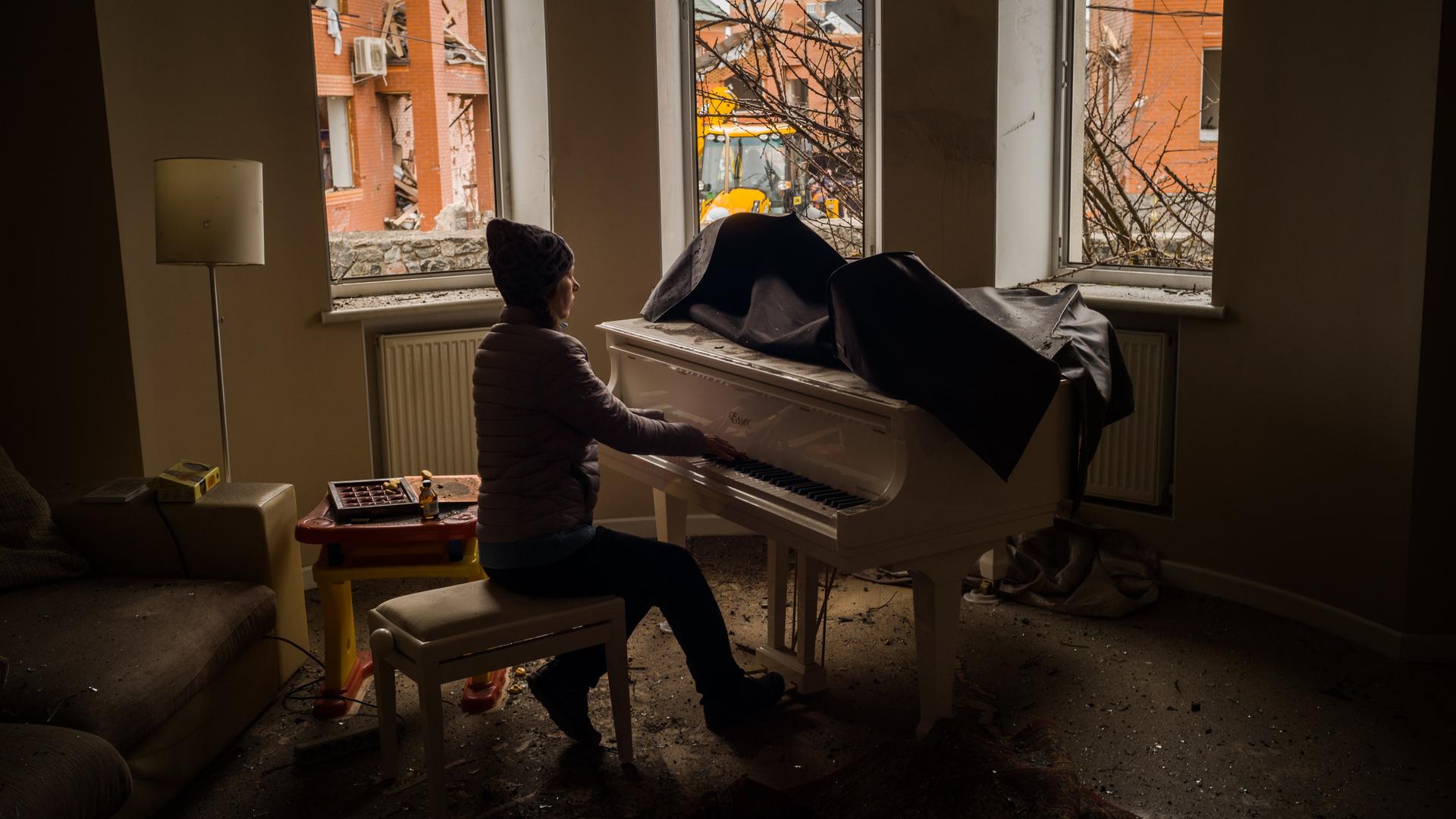 Eine junge Frau spielt Klavier. Durch die zersprungenen Fenster ihrer Wohnung sind im Hintergrund zerstörte Gebäude in der Nachbarschaft zu sehen. Eine junge Frau spielt Klavier. Durch die zersprungenen Fenster ihrer Wohnung sind im Hintergrund zerstörte Gebäude in der Nachbarschaft zu sehen.