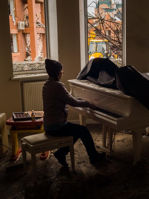 Eine junge Frau spielt Klavier. Durch die zersprungenen Fenster ihrer Wohnung sind im Hintergrund zerstörte Gebäude in der Nachbarschaft zu sehen.
