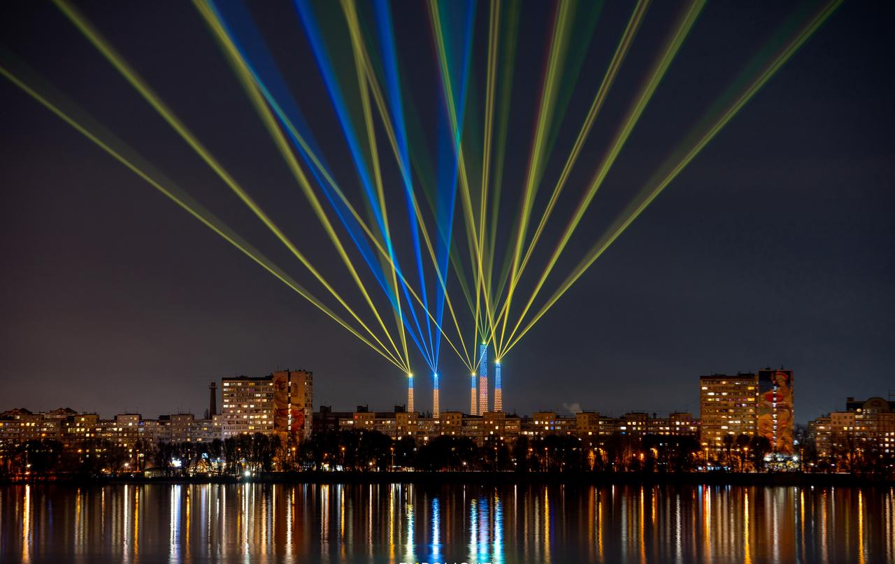 Blaue und gelbe Laserstrahlen von fünf ehemaligen Industrieschornsteinen kreuzen sich im Himmel über der nächtlichen Skyline von Dnipro, deren Lichter sich im Fluss spiegeln.