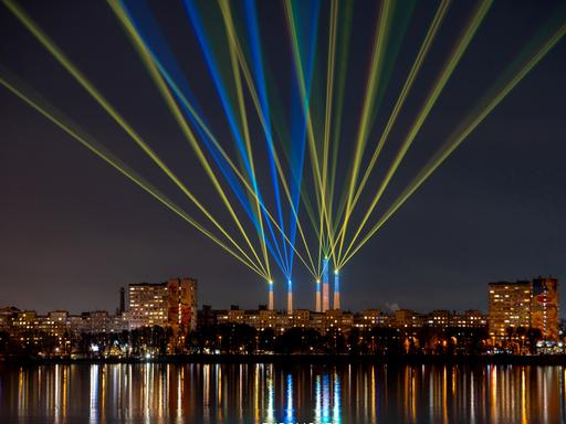 Blaue und gelbe Laserstrahlen von fünf ehemaligen Industrieschornsteinen kreuzen sich im Himmel über der nächtlichen Skyline von Dnipro, deren Lichter sich im Fluss spiegeln. Blaue und gelbe Laserstrahlen von fünf ehemaligen Industrieschornsteinen kreuzen sich im Himmel über der nächtlichen Skyline von Dnipro, deren Lichter sich im Fluss spiegeln.