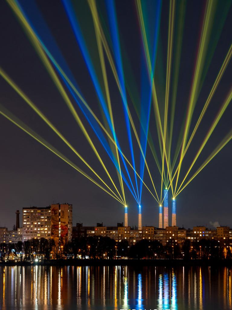 Blaue und gelbe Laserstrahlen von fünf ehemaligen Industrieschornsteinen kreuzen sich im Himmel über der nächtlichen Skyline von Dnipro, deren Lichter sich im Fluss spiegeln. Blaue und gelbe Laserstrahlen von fünf ehemaligen Industrieschornsteinen kreuzen sich im Himmel über der nächtlichen Skyline von Dnipro, deren Lichter sich im Fluss spiegeln.