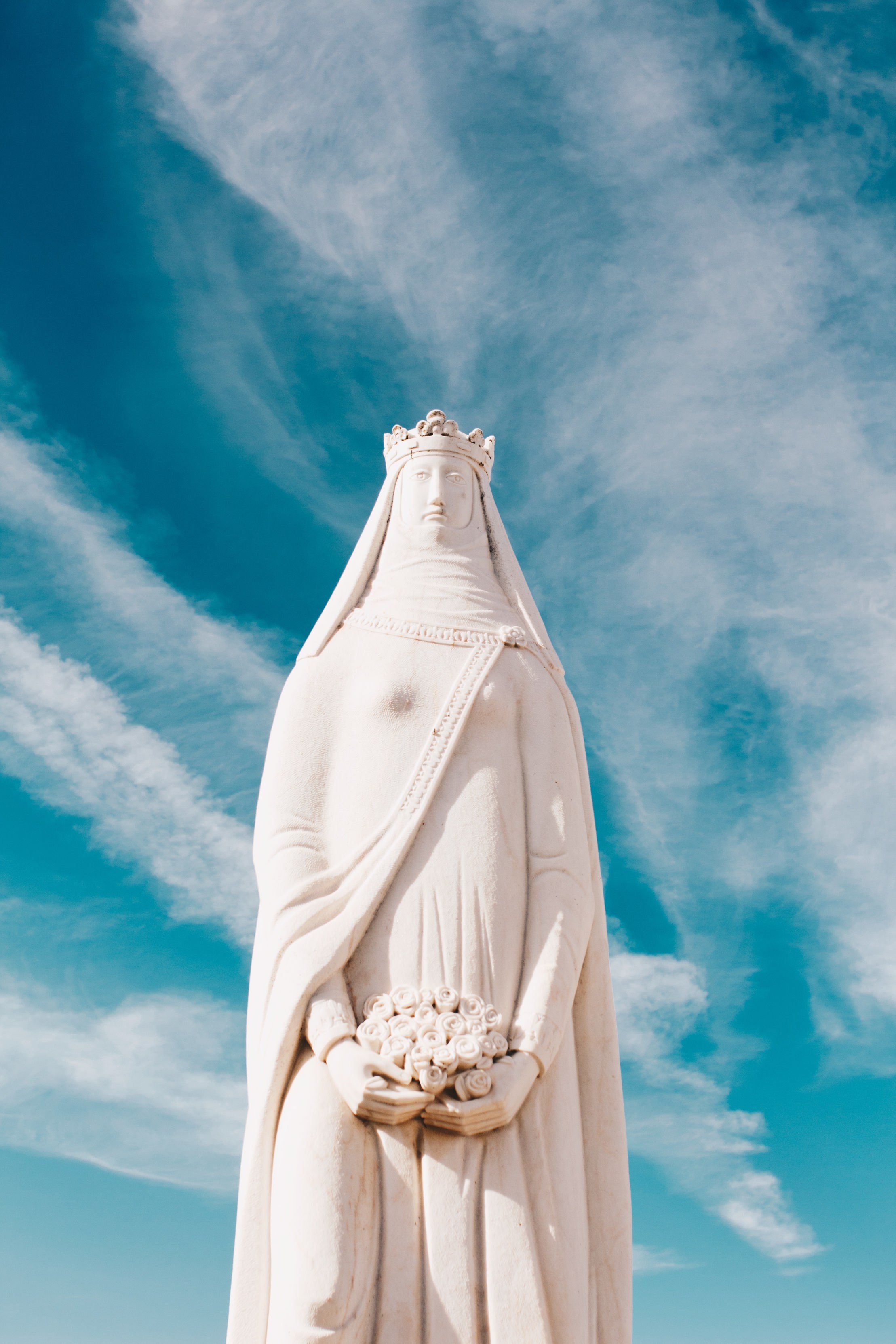 Eine schneeweiße Marienstatue mit Krone steht ernst vor einem blauen Himmel mit interessanten Schleierwolken.