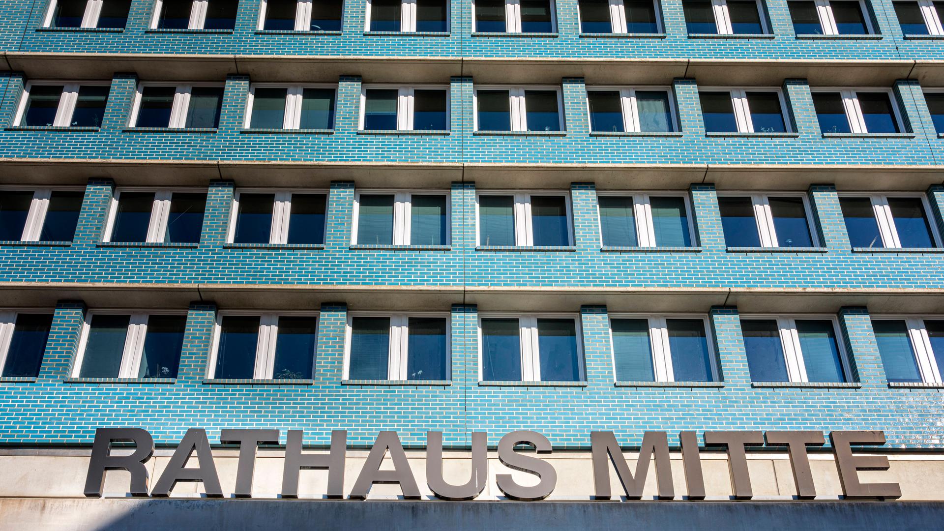 Ein mehrstöckiges Gebäude mit türkisfarbigen Wänden und vielen Fenstern, die gleichförmig angeordnet sind. Im Vordergrund ist der Schriftzug "Rahtahus Mitte" zu sehen, der auf dem Vordach des Gebäudes steht. Ein mehrstöckiges Gebäude mit türkisfarbigen Wänden und vielen Fenstern, die gleichförmig angeordnet sind. Im Vordergrund ist der Schriftzug "Rahtahus Mitte" zu sehen, der auf dem Vordach des Gebäudes steht.