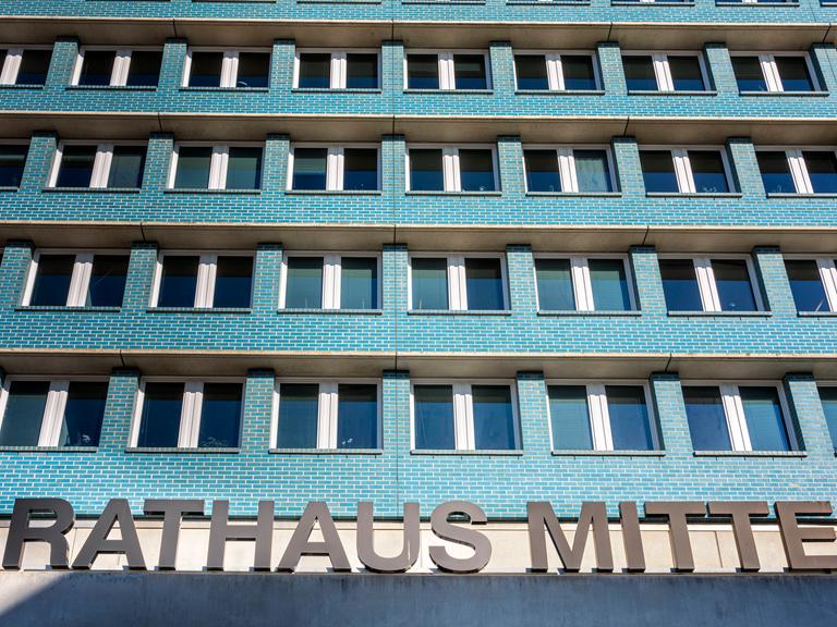 Ein mehrstöckiges Gebäude mit türkisfarbigen Wänden und vielen Fenstern, die gleichförmig angeordnet sind. Im Vordergrund   ist der Schriftzug "Rahtahus Mitte" zu sehen, der auf dem Vordach des Gebäudes steht. 