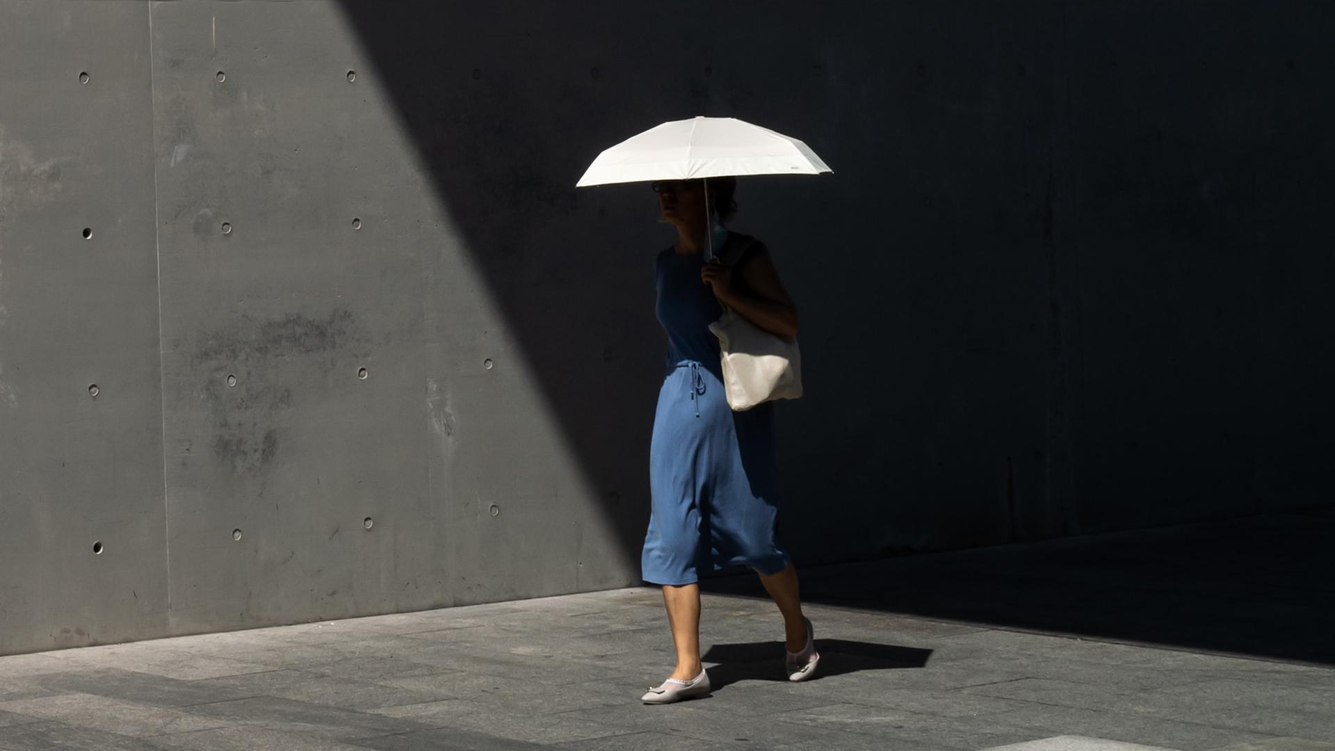 Frau im blauen Kleid, mit weißer Umhängetasche und weißen Schuhen trägt eine weißen Schirm. Sie läuft aus dem Schatten heraus in die Sonne. Gehweg und Tunnelwand sind trist und grau. Ihr Oberkörper ist durch Schatten kaum zu erkennen.