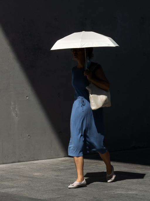 Frau im blauen Kleid, mit weißer Umhängetasche und weißen Schuhen trägt eine weißen Schirm. Sie läuft aus dem Schatten heraus in die Sonne. Gehweg und Tunnelwand sind trist und grau. Ihr Oberkörper ist durch Schatten kaum zu erkennen.