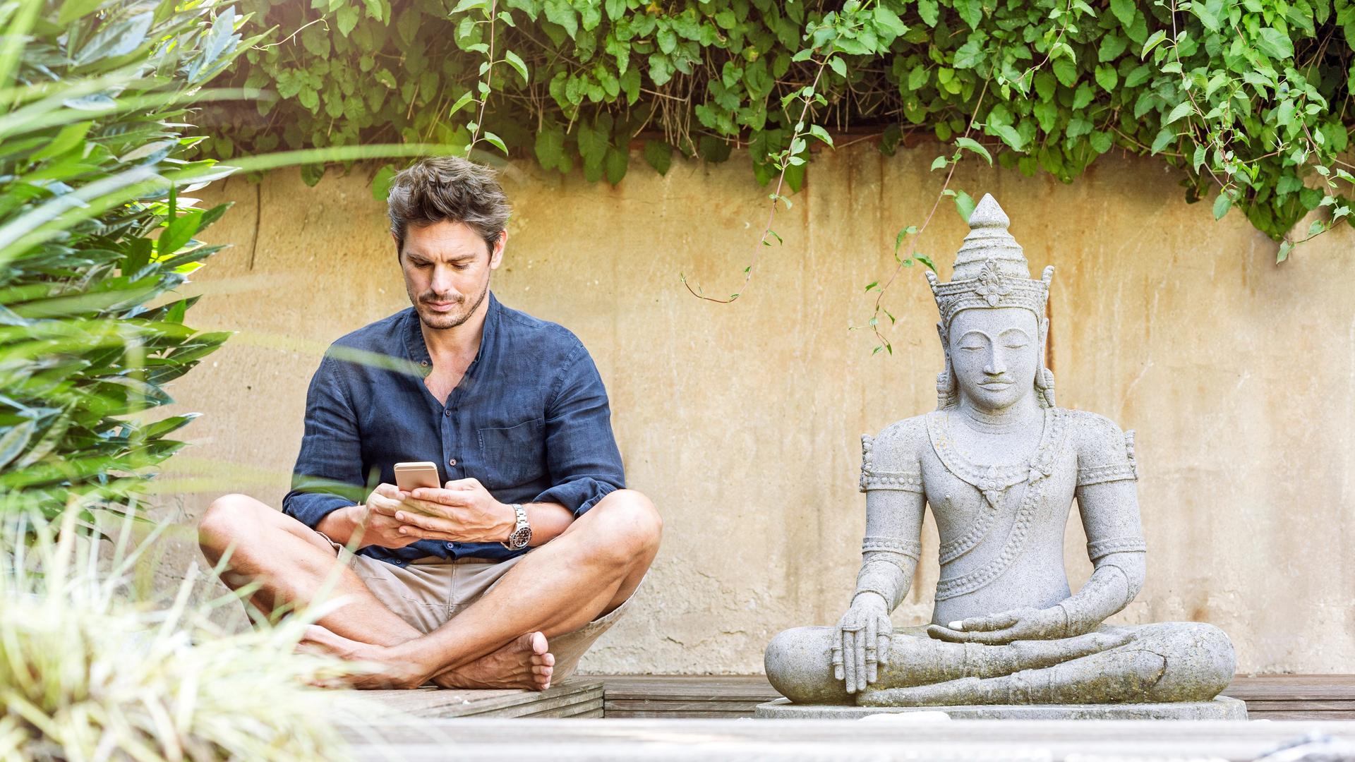 Ein Mann sitzt in einem Zen-Garten im Schneidersitz neben einer Buddhastatue und schaut auf sein Smartphone.