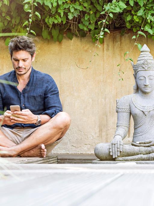 Ein Mann sitzt in einem Zen-Garten im Schneidersitz neben einer Buddhastatue und schaut auf sein Smartphone.