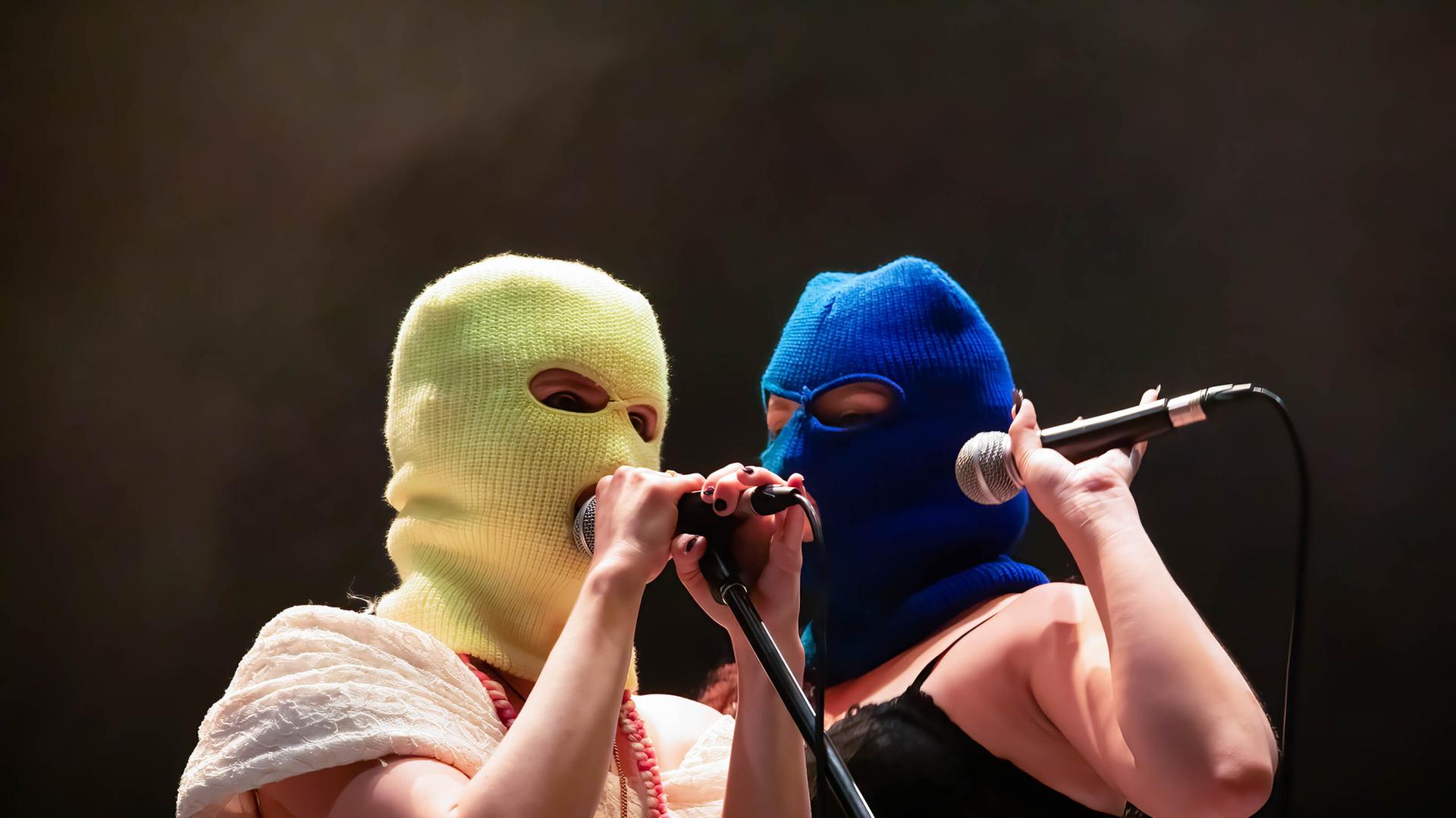 Zwei Frauen stehen auf einer Bühnen und halten ein Mikro in der Hand. Ihre Gesichter kann man nicht erkennen, da sie eine gelbe beziehungsweise blaue Sturmhaube über den Kopf gezogen haben.