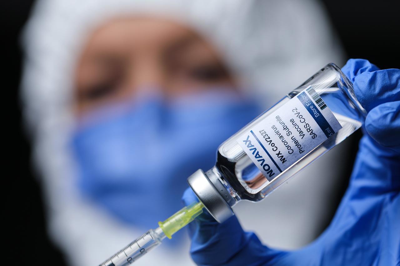 Eine Person in medizinischem Schutzanzug hält ein Fläschchen mit der Aufschrift "Novavax". Eine Person in medizinischem Schutzanzug hält ein Fläschchen mit der Aufschrift "Novavax".
