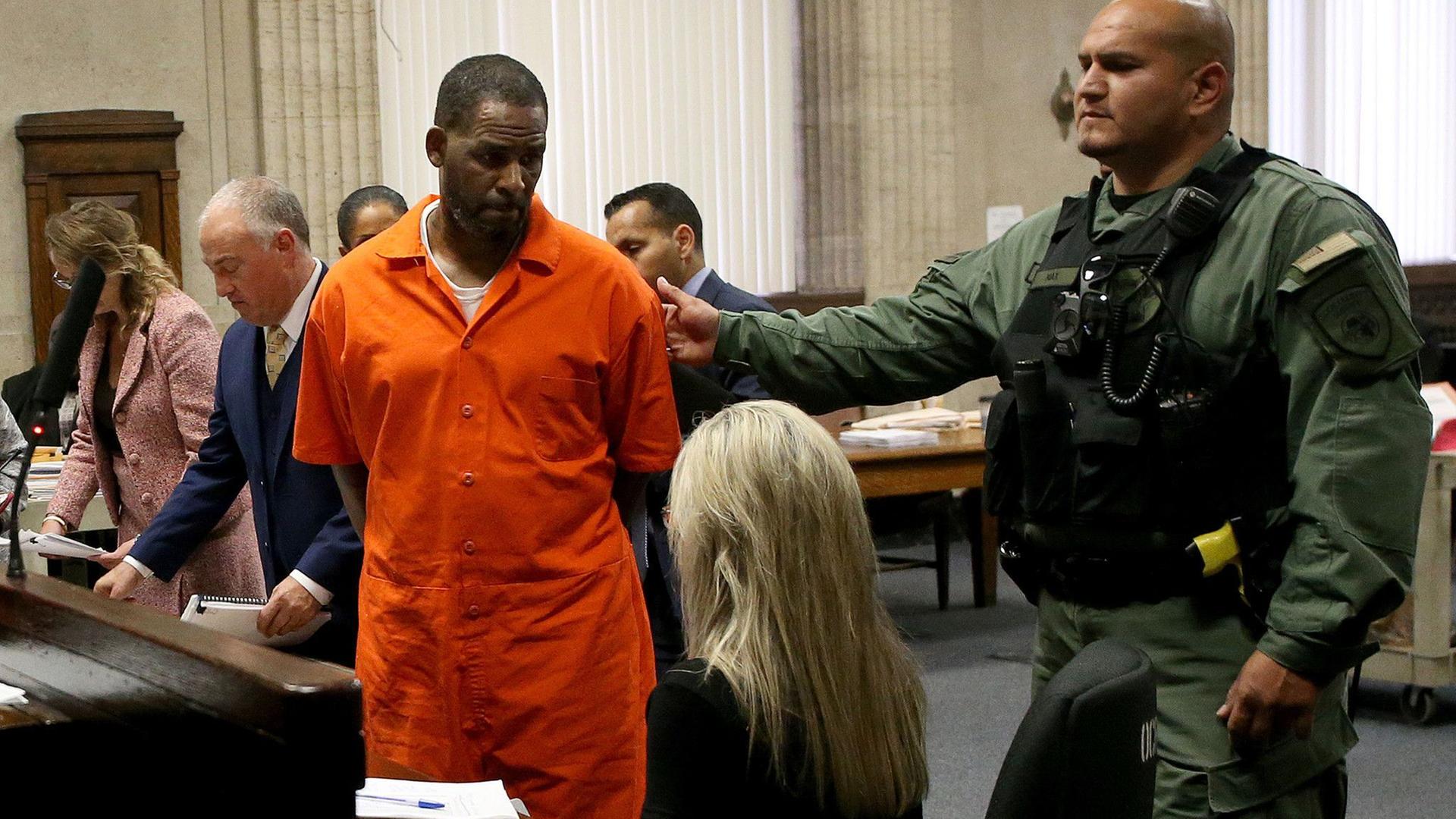 R. Kelly in einem orangen Ganzkörperanzug vor Gericht. Ein Sicherheitsbeamter in Kampfanzug und schusssicherer Weste streckt seine Hand nach ihm aus.