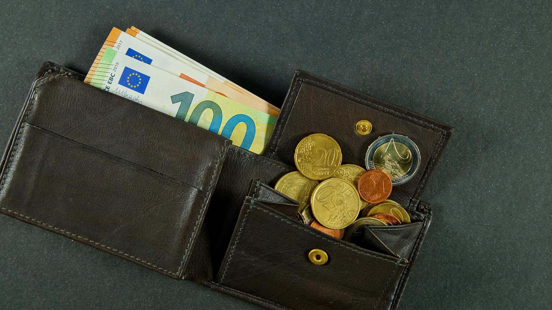 Eine dunkle, geöffnete Ledergeldbörse. Einige Euroscheine sind herausgezogen, Münzen sind aus dem geöffneten Münzfach gerutscht. Eine dunkle, geöffnete Ledergeldbörse. Einige Euroscheine sind herausgezogen, Münzen sind aus dem geöffneten Münzfach gerutscht.