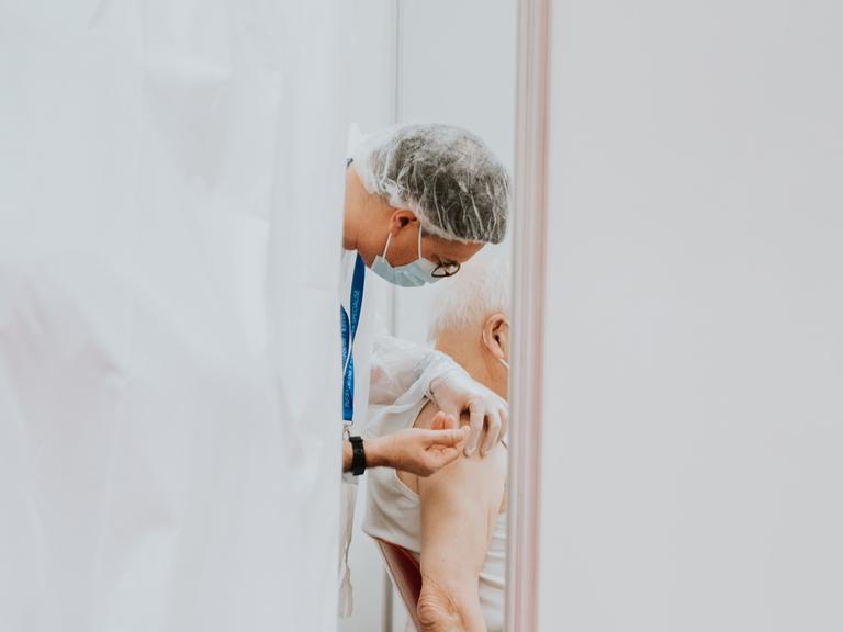 Ein älterer, weißhaariger Mann wird, hinter einem Vorhang, von einem Arzt mit Mundschutz in die Schulter geimpft.