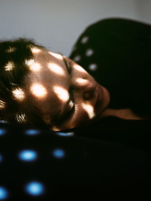 Eine Frau liegt in Fötusstellung auf einem Bett, Sonnensprenkel fallen durch eine halb geschlossene Jalousie auf sie.