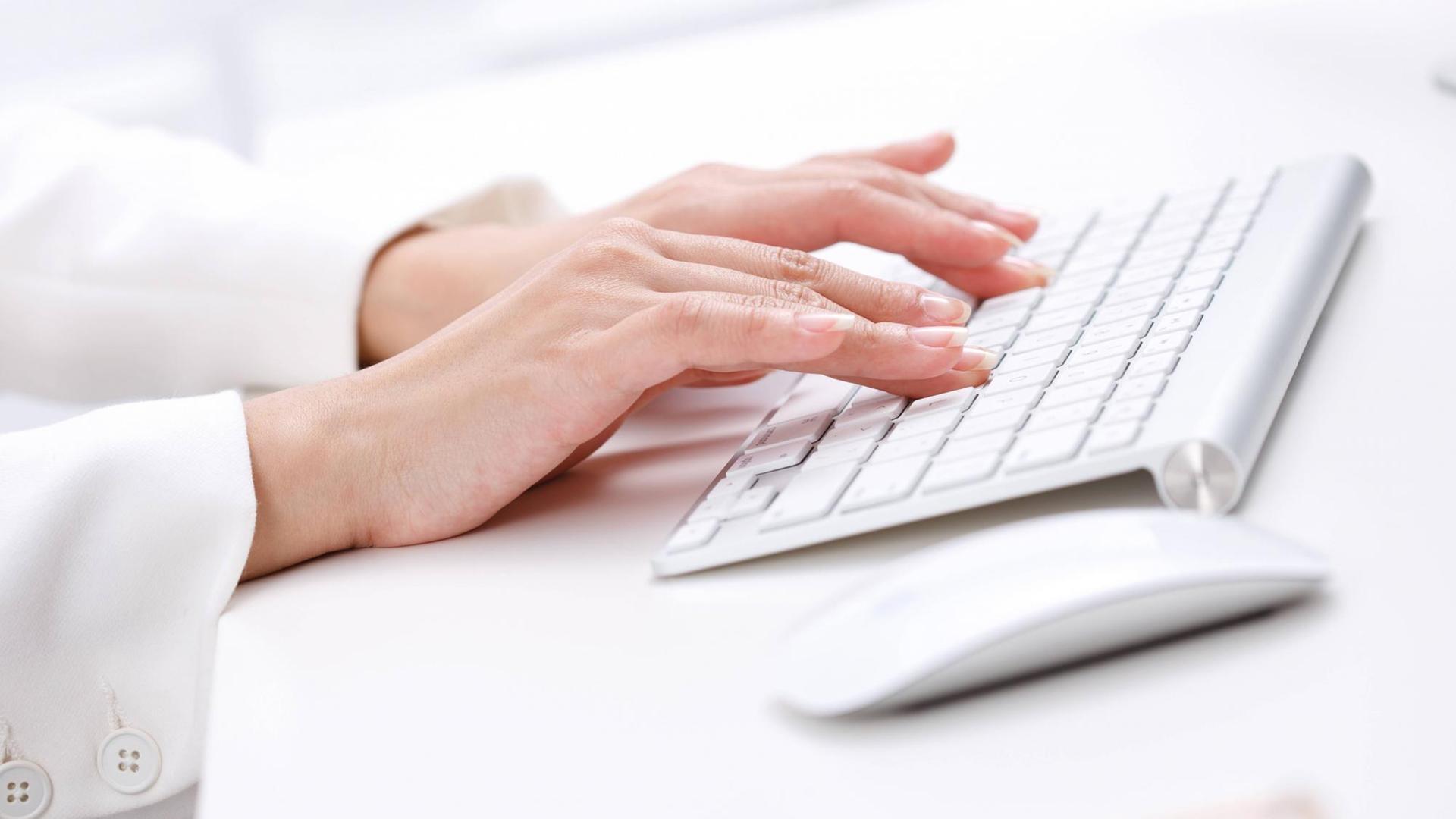 Frauenhände tippen auf einer weißen Computertastatur. Rechts daneben liegt eine weiße Computermaus. Frauenhände tippen auf einer weißen Computertastatur. Rechts daneben liegt eine weiße Computermaus.