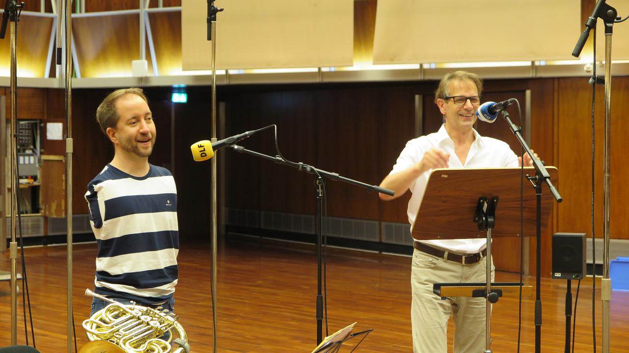 Ein Hornist ohne Arme wird in einem holzgetäfelten Raum mit Mikrofonen von einem Journalisten interviewt. Danaben liegt auf einem Stuhl sein Instrument. Ein Hornist ohne Arme wird in einem holzgetäfelten Raum mit Mikrofonen von einem Journalisten interviewt. Danaben liegt auf einem Stuhl sein Instrument.