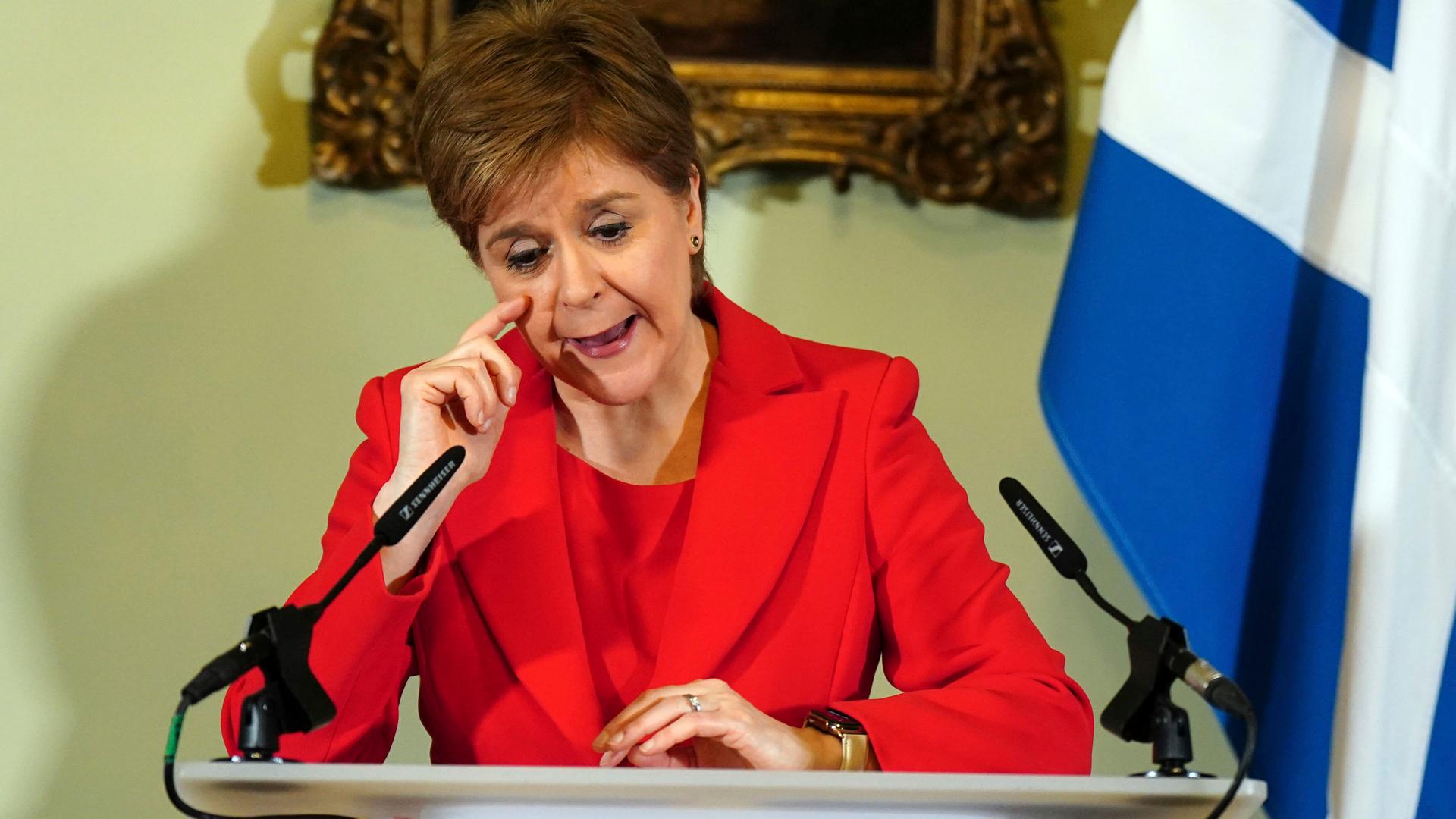 Nicola Sturgeon trägt einen roten Anzug. Sie steht hinter einem Pult und redet. Nicola Sturgeon trägt einen roten Anzug. Sie steht hinter einem Pult und redet.