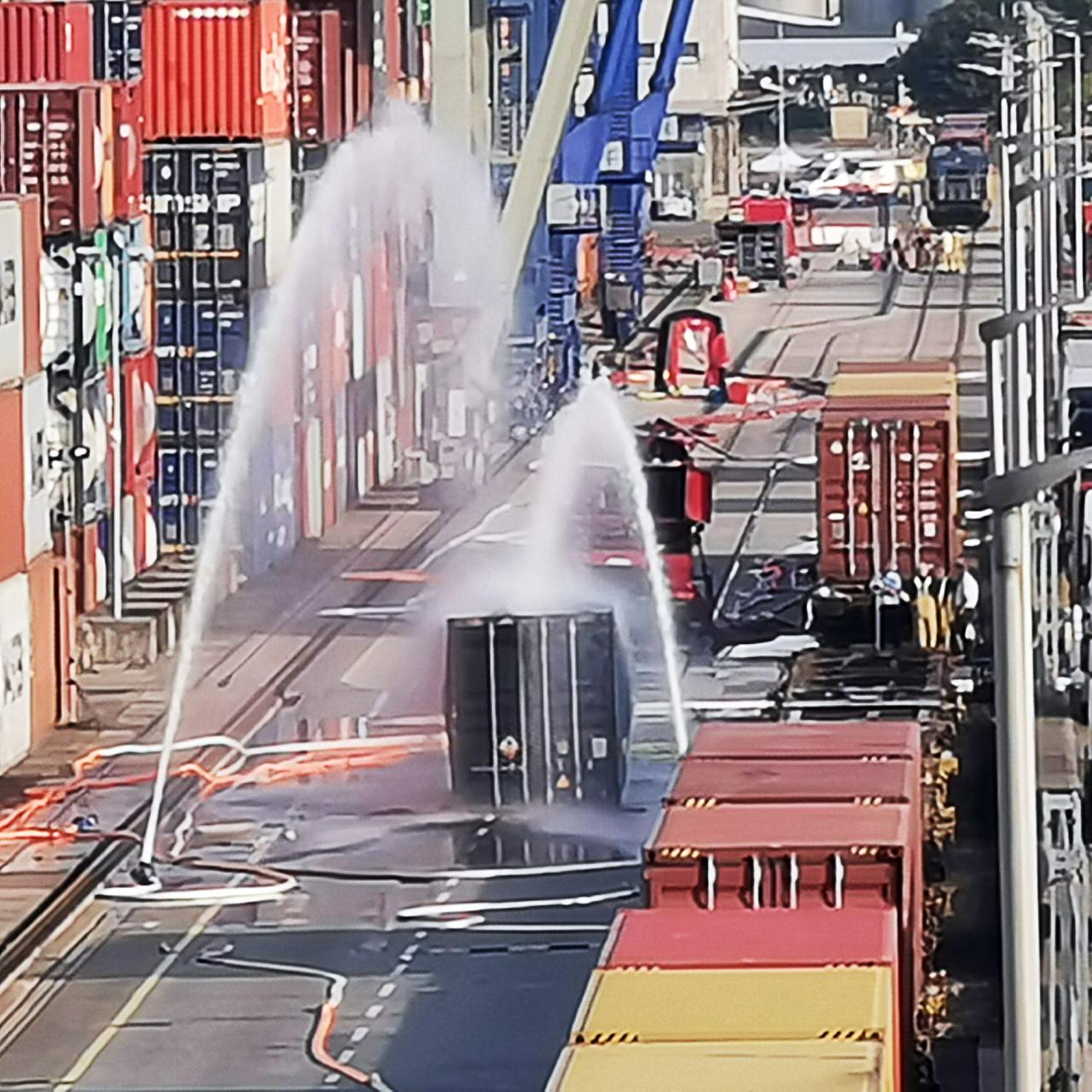Wasserfontänen benetzen einen Seecontainer auf einem Hafengelände.