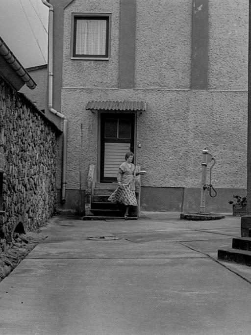 Fotografie einer Frau mit einem Teller in der Hand, die ein Haus in Richtung Hof in Feldberg im Jahr 1989 verlässt.