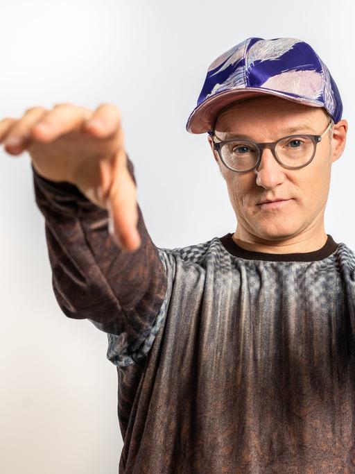 Der Musiker PeterLicht mit Kappe steht der Kamera gegenüber und streckt dabei Arme und Hände nach ihr aus.