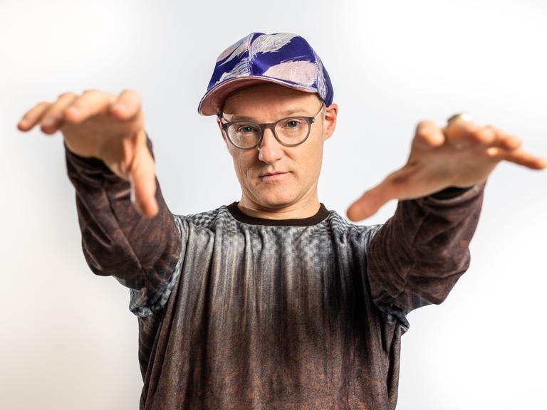 Der Musiker PeterLicht mit Kappe steht der Kamera gegenüber und streckt dabei Arme und Hände nach ihr aus.