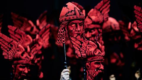 Auf der Bühne halten Darsteller bei der "Götterdämmerung" rote Masken in die Höhe.