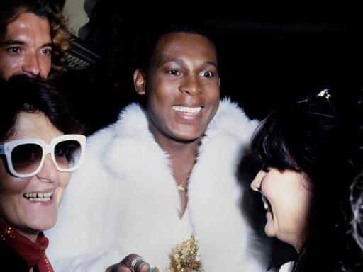 Der Discosänger Sylvester trägt einen weißen Pelzmantel und gibt Fans lachend Autogramme