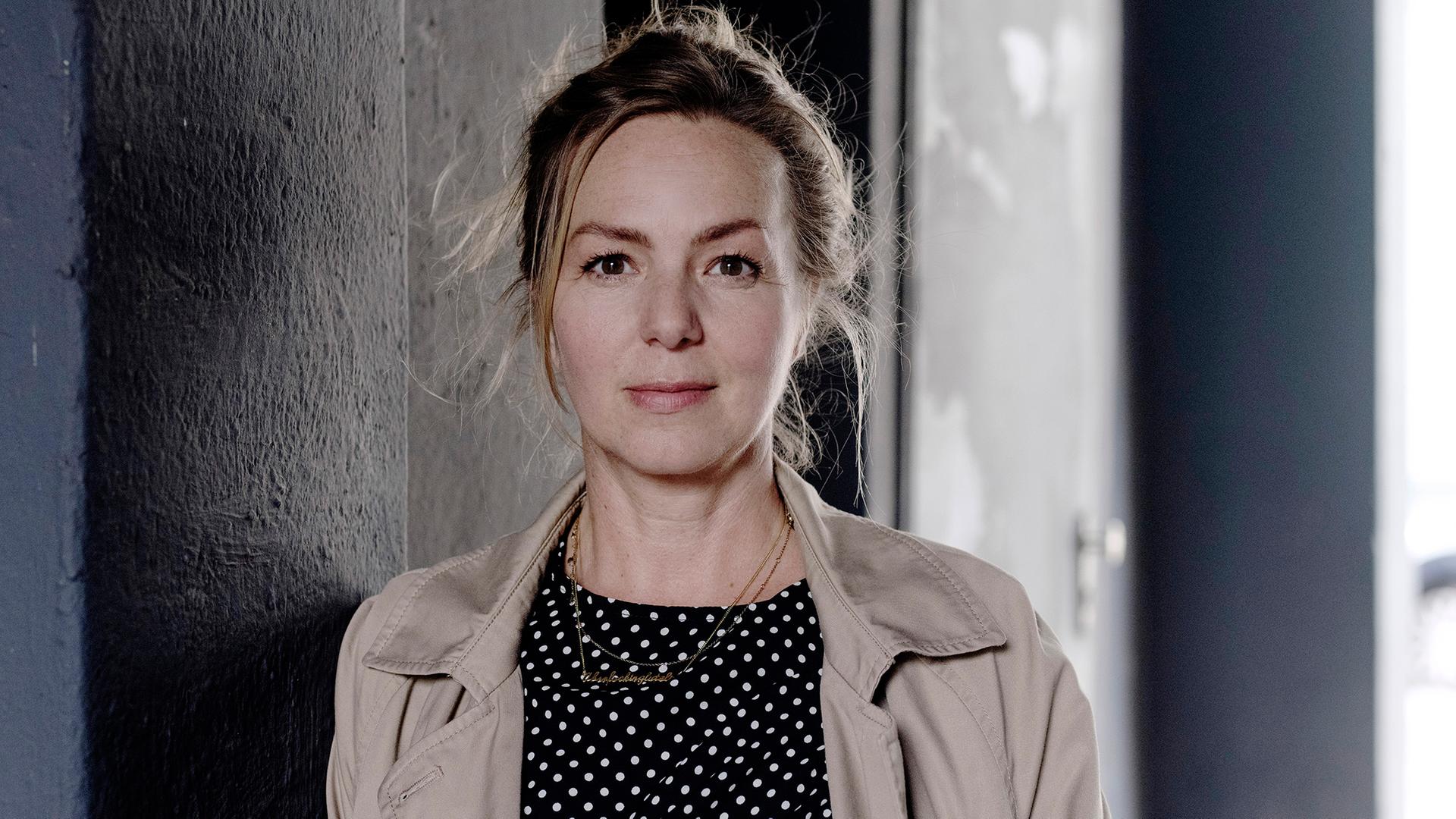 Simone Buchholz schaut in die Kamera. Sie trägt einen Trenchcoat und darunter ein Oberteil in dunkler Farbe mit weißen Punkten. Ihre langen Haare hat sie hochgesteckt. Simone Buchholz schaut in die Kamera. Sie trägt einen Trenchcoat und darunter ein Oberteil in dunkler Farbe mit weißen Punkten. Ihre langen Haare hat sie hochgesteckt.
